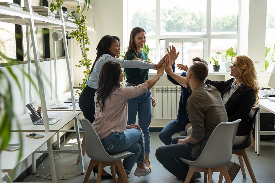 Employee Benefits - Group Of Employees Sitting In Office Giving Eachother High Fives